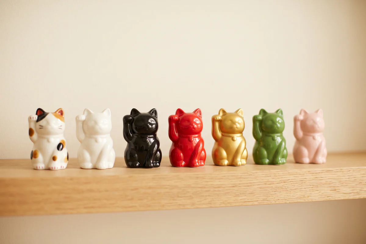 Sept maneki-neko en céramique, patte droite levée, alignés sur une étagère : tricolore, blanc, noir, rouge, doré, vert et rose